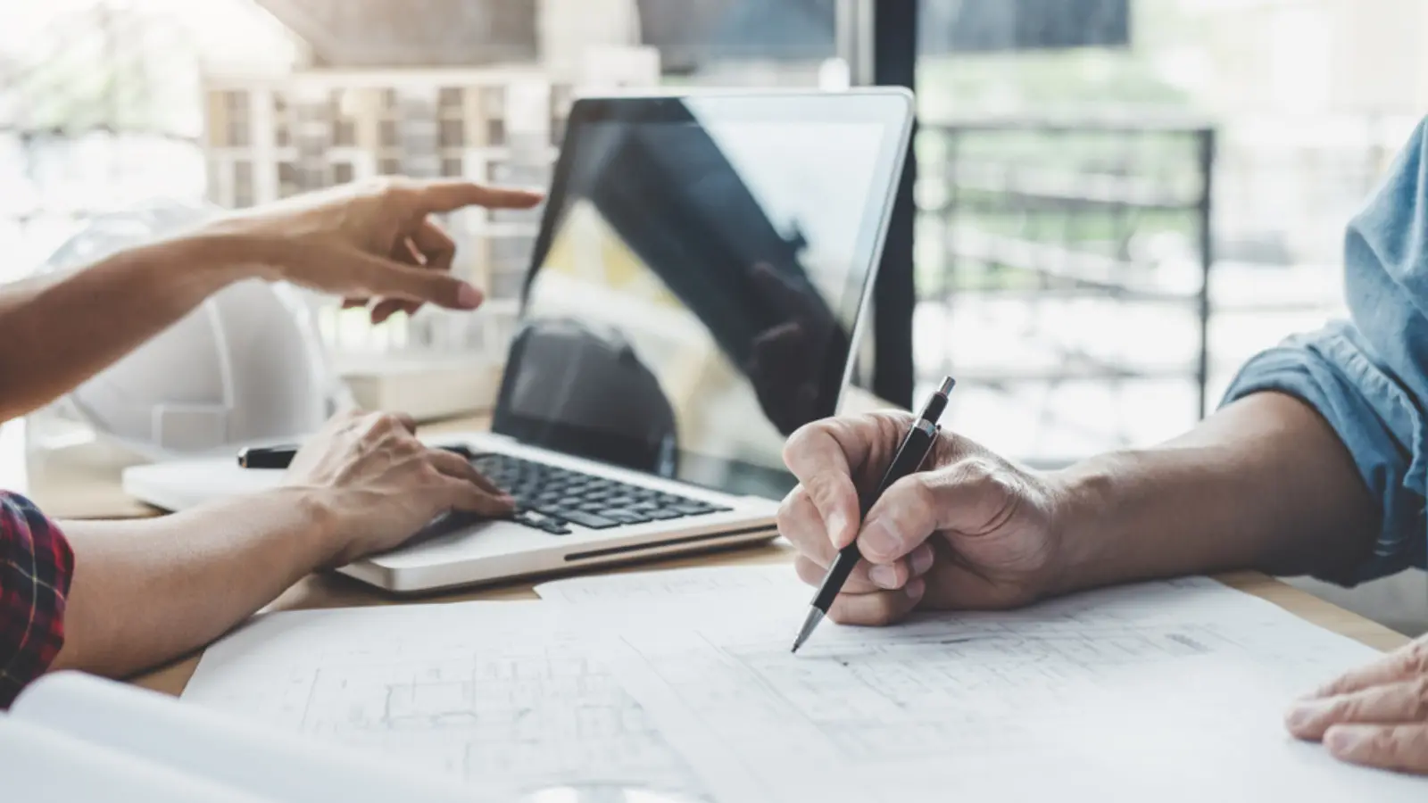 Project manager reviewing cost plans, drawings and a laptop with floor plans spread across a meeting table.