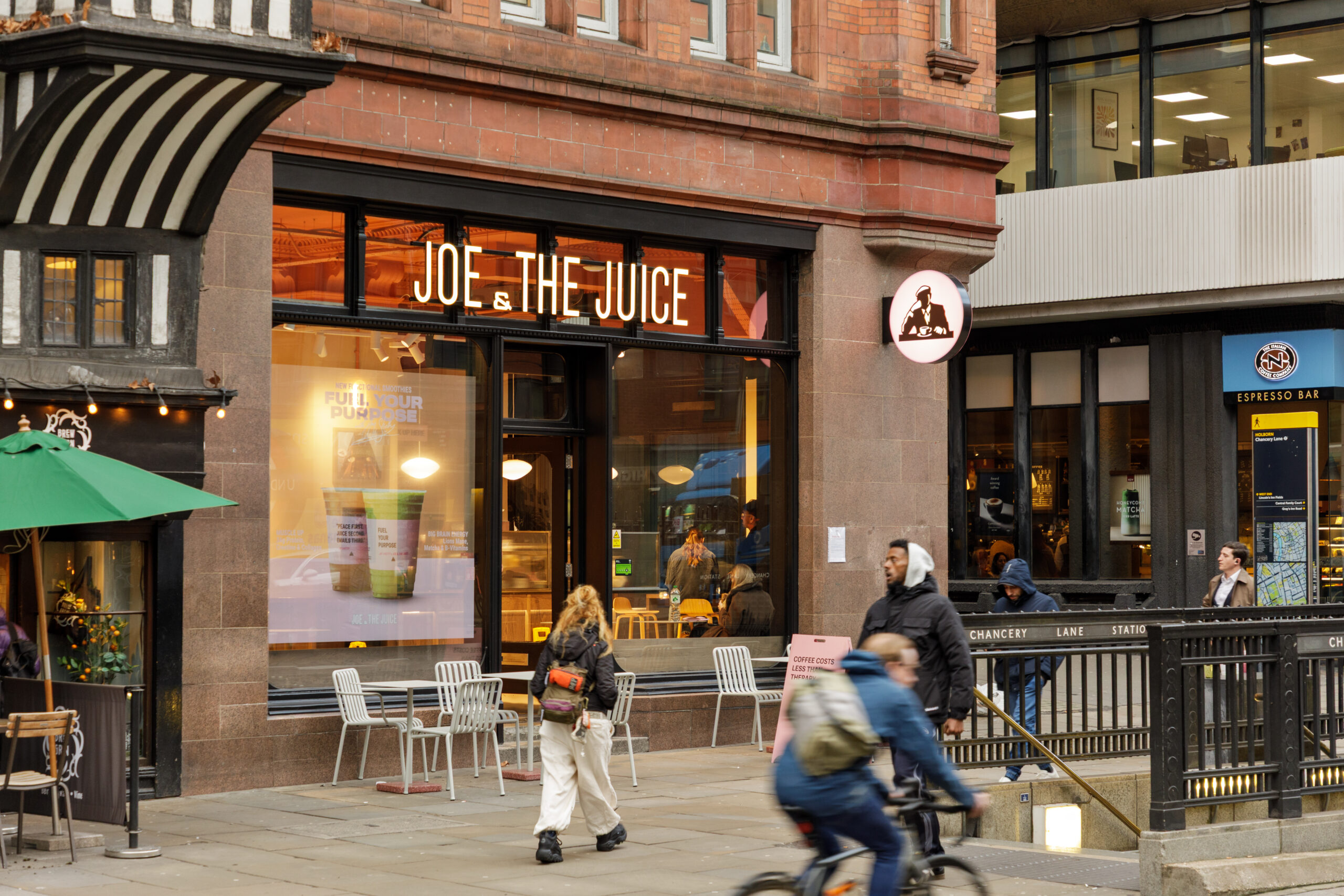 Joe & The Juice illuminated shopfront on a red-brick High Holborn building, with pavement seating and Chancery Lane station entrance alongside.